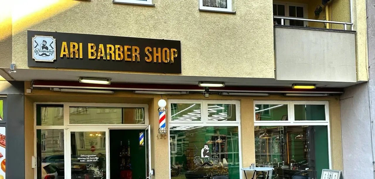 Außenansicht von Ari Barbershop mit goldenem Schriftzug, Barber-Pole und grünem Innenraum hinter den Fenstern